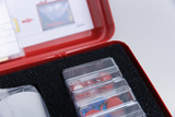 Das Bild zeigt eine rote Aufbewahrungsbox, die verschiedene Experimentiermaterialien enthält, darunter farbige Schaumstoffscheiben und nummerierte Plättchen in durchsichtigen Fächern. Im Hintergrund ist eine Anleitung oder ein Hinweisblatt zu sehen, das m