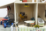 Das Bild zeigt ein Modell eines Hauses, das als Lehrmittel im Experimentierbedarf verwendet wird. Im Inneren sind ein Wohnzimmer mit Möbeln, einem Fernseher und einer Küche zu sehen, sowie technische Komponenten für Elektronikexperimente.