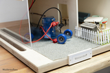 Das Bild zeigt ein Modell, das eine kleine Fahrzeugkonstruktion mit blauen Rädern und einem transparenten Motor im Vordergrund darstellt. Im Hintergrund sind eine Miniaturumgebung mit Gartenzaun, Mülleimer und Möbeln zu sehen, das Thema des Modells schein