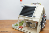 Das Bild zeigt ein Modell eines Hauses mit einem Solarpanel auf dem Dach und verschiedenen experimentellen Geräten, die zur Demonstration von Energieumwandlung und -nutzung konzipiert sind. Eine kleine Gartenlandschaft mit Zäunen und Dekoration rundet die