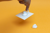 Das Bild zeigt eine Hand, die ein weißes Experimentiergerät mit einem Saugnapf hält, während ein weiterer Saugnapf daneben liegt. Der Hintergrund ist in einem leuchtenden Orange gehalten, was das Augenmerk auf das Experimentiermaterial lenkt.