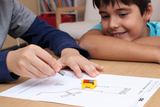 Das Bild zeigt zwei Kinder, die aktiv an einem Experimentierprojekt arbeiten. Ein Junge hebt einen Magneten über ein Papier mit einer gezeichneten Straße, während der andere gespannt zuschaut und ein kleines gelbes Fahrzeug in der Mitte platziert ist.