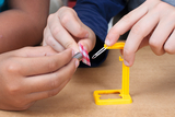 Auf dem Bild sind zwei Kinderhände zu sehen, die an einem Experimentiergerät arbeiten. Eines der Kinder hält einen kleinen metallischen Gegenstand, während das andere eine rot-weiß karierte Stoffbahn mit einer gelben Klammer stabilisiert.