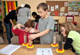 Auf dem Bild experimentieren Kinder in einem Klassenzimmer mit verschiedenen Materialien in durchsichtigen Behältern. Sie fügen Wasser zu den Behältern hinzu und beobachten die Ergebnisse, wobei ihre Neugier und Zusammenarbeit deutlich sichtbar sind.