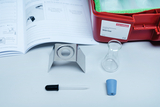 Das Bild zeigt einen Experimentierbedarf mit einem Anleitungshandbuch, einem Stativ, einem Erlenmeyerkolben, einer Pipette und einem blauen Stopfen. Diese Materialien sind für Experimente zum Thema Wärme im Bildungsbereich gedacht.