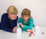 Das Bild zeigt zwei Kinder, die an einem Experimentierplatz sitzen und konzentriert mit verschiedenen Laborgeräten arbeiten. Eine rot gefärbte Flüssigkeit in einem Erlenmeyer geht im Vordergrund auf, während die Kinder eine Pipette und einen Messzylinder 