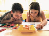 Das Bild zeigt zwei Kinder, die an einem Tisch sitzen und gespannt ein Experiment mit einer Glaskaraffe beobachten, die Wasser und bunte Figuren enthält. Um sie herum liegen weitere Experimentiermaterialien und Notizblätter, was auf eine Lerngeschichte im