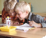 Das Bild zeigt zwei Kinder, die neugierig ein Experiment durchführen. Vor ihnen steht ein Gerät mit einer gelben Kugel, während sie konzentriert auf ein Blatt Papier mit Anweisungen schauen.