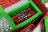 Das Bild zeigt eine rote Aufbewahrungsbox mit grünem Schaumstoff, in dem mehrere durchsichtige, rote Versuchsröhrchen gelagert sind. Zusätzlich sind kleine Fächer im oberen Bereich der Box sichtbar, die diverse Zubehörteile enthalten.