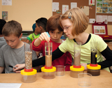 Das Bild zeigt Kinder, die in einem Klassenzimmer mit verschiedenen Behältern experimentieren. Sie führen offenbar ein Experiment durch, bei dem sie verschiedene Materialien in durchsichtige Zylinder schichten und beobachten.