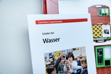 Das Bild zeigt ein Schüler-Set zum Experimentieren mit Wasser, präsentiert von Cornelsen Experimenta. Im Vordergrund sind Kinder zu sehen, die gemeinsam verschiedene Experimente durchführen und dabei Neugier und Teamarbeit demonstrieren.