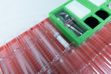 Das Bild zeigt eine Anordnung von Reagenzgläsern in einem roten Halter sowie ein grünes Set mit Laborutensilien, einschließlich Pipetten und Platten. Im Vordergrund befindet sich ein Behälter mit einer Kennzeichnung für Chemikalien.