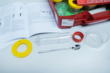 Das Bild zeigt ein Experimentier-Set mit verschiedenen Werkzeugen, darunter ein transparentes Röhrchen, ein dazugehöri­ges Behältnis und eine Löffel. Außerdem liegt ein Handbuch auf, das Anweisungen für die Durchführung von Experimenten enthält.