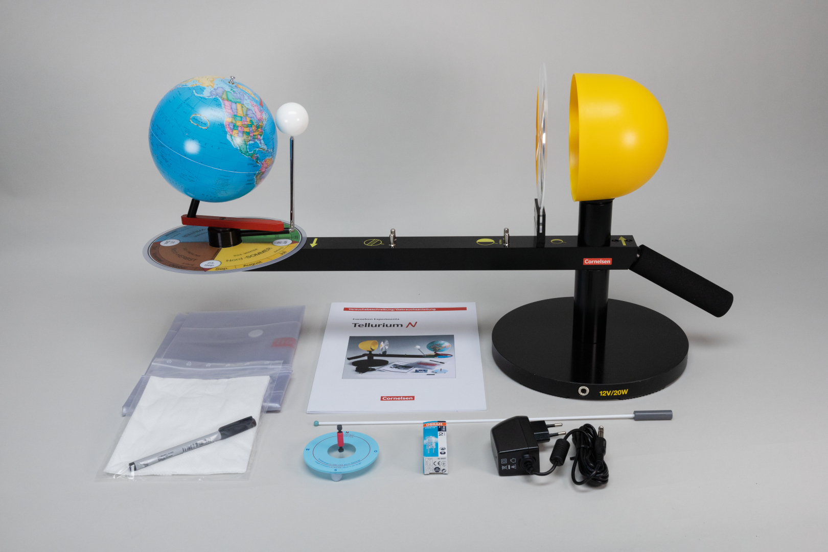 Das Bild zeigt ein physikalisches Experimentierkit mit einer Weltkugel, einer gelben Halbkugel und verschiedenen Zubehörteilen wie einer Anleitung, Kabeln und Messinstrumenten. Es dient zur Veranschaulichung von Konzepten wie Lichtstrahlung und Tag-Nacht-