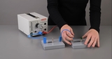 Das Bild zeigt eine Experimentierstation mit einer Stromversorgungseinheit und zwei Schaltungen, die mit Drähten verbunden sind. Eine Person führt eine Verbindung zu einer Lampe her, um elektrische Schaltungen zu demonstrieren.