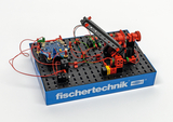 Das Bild zeigt ein fischertechnik-Modell, das aus einer Basisplatte und verschiedenen Konstruktionselementen besteht, darunter elektronische Bauteile und Kabel. Es könnte für Experimente im Bereich Technik und Robotik im Bildungswesen verwendet werden.