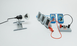 Das Bild zeigt eine elektrische Lampe, die auf einem Ständer montiert ist, sowie mehrere Messgeräte und Versuchsanordnungen auf einer Arbeitsfläche. Die Geräte sind miteinander verbunden, um Experimente im Bereich Elektrizität und Messtechnik durchzuführe