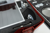 Das Bild zeigt eine aus Kunststoff gefertigte Box, die mit Schaumstoffpolstern gefüllt ist und mehrere experimental-technische Komponenten enthält. Zu sehen sind ein transparenter Deckel, metallische Teile und andere Objekte, die für Experimente im Bildun