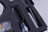 Das Bild zeigt eine sortierte Werkzeugbox mit verschiedenen Experimentierutensilien, darunter Glasröhren und einen runden, spiegelnden Behälter. Die Utensilien sind ordentlich in einem schaumstoffgepolsterten Fach angeordnet, was auf eine sorgfältige Präsentation hinweist.