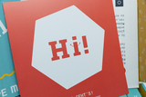 Das Bild zeigt eine bunte Ansammlung von Broschüren und Anleitungen aus einem Online-Shop für Experimentierbedarf. Besonders hervorgehoben ist eine rote Karte mit dem fröhlichen Schriftzug "Hi!" in der Mitte.