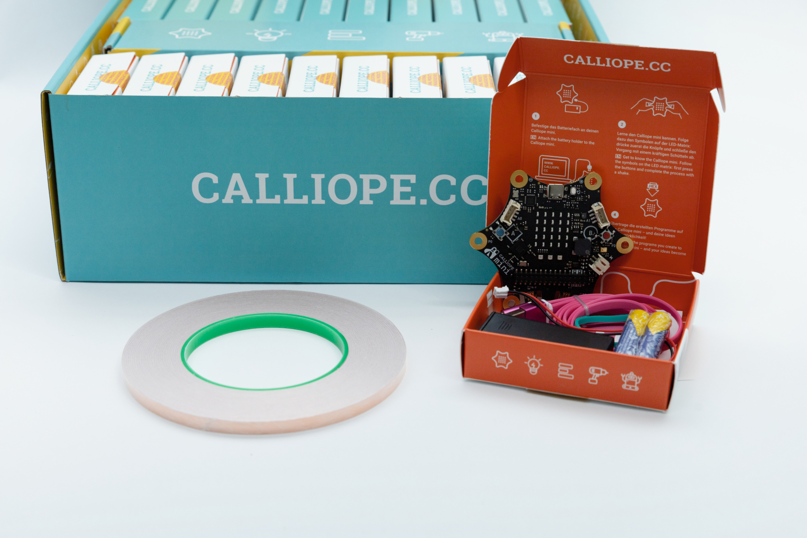 Das Bild zeigt ein Calliope mini, ein Mikrocontroller-Board, das zur kreativen Programmierung und zum Experimentieren in der Bildung verwendet wird. Neben dem Board sind pinke Kabel und eine Rolle Klebeband auf einem neutralen Hintergrund zu sehen.