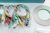 Das Bild zeigt mehrere Packungen mit bunten Drahtverbindungen, die jeweils verschiedenen Farben zugeordnet sind, sowie eine Rolle Klebeband und eine kleine Tüte mit LEDs. Diese Materialien eignen sich für Experimente in Bildungseinrichtungen.