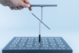 Das Bild zeigt eine Hand, die ein Experimentiergerät hält, das aus einer vertikalen Stange mit einem beweglichen Element besteht. Das Gerät ist auf einer unterteilten, flachen Basis platziert, die zahlreiche Anschlussmöglichkeiten bietet und für experimentelle Zwecke im Bildungsbereich verwendet wird.
