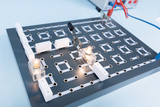 Das Bild zeigt eine Experimentierplatine mit verschiedenen Anschlussmöglichkeiten, auf der zwei Glühbirnen leuchten. Angeklemmt sind zwei Kabel, die eine elektrische Verbindung herstellen, und die Anordnung lässt sich für verschiedene Experimente im Bildungsbereich nutzen.