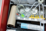 Das Bild zeigt eine Sammlung von Experimentiermaterialien, darunter verschiedene Bechergläser, Pipetten und ein thermisches Gerät. Diese Materialien sind geordnet in einem roten Kasten, der für den Einsatz im Bildungsbereich konzipiert ist.