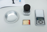 Das Bild zeigt verschiedene Materialien für Experimente, darunter eine Aluminium-Schale, Eisenwolle in einem Behälter, ein Paket mit Sicherheitsstreichhölzern und eine digitale Waage. Diese Utensilien sind wahrscheinlich für chemische oder physikalische Versuche im Bildungsbereich gedacht.