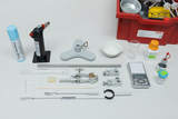 Das Bild zeigt eine Sammlung von Experimentiermaterialien, darunter ein Bunsenbrenner, Reagenzgläser, eine elektronische Waage und verschiedene chemische Substanzen. Die Utensilien sind für den Einsatz im Bildungsbereich konzipiert und in einer roten Aufbewahrungsbox organisiert.