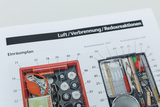 Das Bild zeigt eine detaillierte Auflistung der Inhalte eines Experimentierkits für das Thema Luft, Verbrennung und Redoxreaktionen. Jedes Item ist mit einer Nummer versehen, die auf einen Plan zur Anordnung der Materialien in einer rot-schwarzen Kunststoffbox verweist.