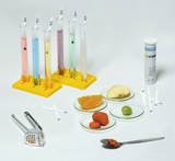 Das Bild zeigt einen Experimentieraufbau mit fünf Reagenzgläsern, die jeweils gefärbte Flüssigkeiten enthalten, sowie verschiedenen Obststücken auf kleinen Petri-Schalen. Neben dem Obst liegen pH-Teststreifen und ein Küchengerät, vermutlich zur Verarbeitung der Lebensmittel.