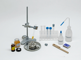 Das Bild zeigt eine Sammlung von Laborgeräten und Chemikalien, darunter Reagenzgläser, eine Bunsenbrenner, Pipetten und verschiedene Flaschen mit Flüssigkeiten. Die Anordnung der Geräte auf einem weißen Hintergrund vermittelt einen aufgeräumten, professionellen Eindruck.