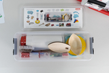 Das Bild zeigt eine transparente Box mit verschiedenen Experimentiermaterialien, darunter Holzlöffel, bunte Scheiben und weitere Utensilien. Oben befindet sich eine Anleitung mit Abbildungen der enthaltenen Materialien, die für Bildungszwecke genutzt werden können.