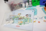 Das Bild zeigt ein Experimentierpaket mit Anleitungen und Materialien für ein Projekt über Wasser. Es enthält eine illustrierte Broschüre sowie verschiedene Utensilien, die für das Experimentieren im Bildungsbereich genutzt werden können.