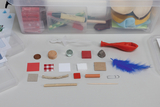 Das Bild zeigt eine Sammlung verschiedener Materialien, darunter Farben, Stoffe, ein Luftballon und andere Objekte, die für Experimente und kreative Projekte im Bildungsbereich verwendet werden können. Die Gegenstände sind ordentlich angeordnet und in einer transparenten Box sichtbar.