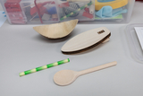 Das Bild zeigt verschiedene experimentelle Materialien, darunter zwei Holzformen, einen Holzlöffel und ein buntes, gestreiftes Rohr. Im Hintergrund sind weitere Bastel- und Experimentiermaterialien in durchsichtigen Boxen sichtbar.