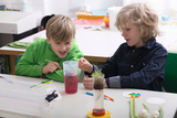 Auf dem Bild sind zwei Kinder zu sehen, die in einem Experimentierraum miteinander arbeiten. Sie untersuchen einen Behälter mit roter Flüssigkeit und einem Glas mit Erde und Pflanzen, während sie gespannt und neugierig interagieren.