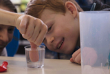 Das Bild zeigt ein Kind, das fasziniert auf ein Taschenspielertrick-Experiment schaut, bei dem ein Finger in ein Glas Wasser eingetaucht wird. Im Hintergrund sind weitere Kinder zu sehen, die ebenfalls interessiert zuschauen.