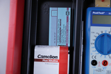 Das Bild zeigt eine Packung mit Lackmuspapier zur pH-Wert-Bestimmung und eine Batterie der Marke Camelion. Außerdem ist ein Multimeter zu sehen, das für verschiedene elektrische Messungen verwendet werden kann.
