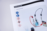 Das Bild zeigt eine Seite eines Lehrbuchs über Elektrochemie, das sich mit dem Thema Galvanisierung beschäftigt. Es sind Sicherheits- und Gefahrenzeichen sowie Abbildungen von Experimentiermaterialien wie einer elektrochemischen Zelle und Zubehör zu sehen.