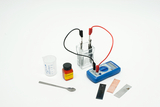 Das Bild zeigt experimentelle Ausrüstung, bestehend aus einem Becherglas mit Flüssigkeit, zwei Elektroden, einem Multimeter zur Messung elektrischer Werte sowie verschiedenen Metallproben und einer Verpackung mit Zitronensäure. Diese Materialien sind für chemische und physikalische Experimente im Bildungswesen geeignet.