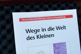 Das Bild zeigt eine Anleitung mit dem Titel „Wege in die Welt des Kleinen“, die wahrscheinlich Experimente für das Bildungswesen beschreibt. Die Unterlage liegt auf einem geschäumten Untergrund, der typischerweise für den Schutz von Materialien verwendet wird.