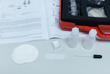 Das Bild zeigt eine Sammlung von Experimentiermaterialien, darunter Reagenzgläser, Pipetten, Objektträger und eine Anleitung, die sich auf biologische Experimente bezieht. Die Materialien sind in einem roten Koffer organisiert, der für den Einsatz im Bildungsbereich konzipiert ist.