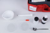 Das Bild zeigt ein Experimentierset für landwirtschaftliche Nutzpflanzen, das in einem roten Kasten verpackt ist. Es enthält verschiedene Laborutensilien wie Glasplatten, eine Pipette, chemische Substanzen und einen Messbecher, die für Versuche verwendet werden können.