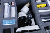 Das Bild zeigt eine kompakte Aufbewahrung für Experimentierbedarf, die ein Mikroskop sowie verschiedene Zubehörteile enthält. Zu den sichtbaren Elementen gehören Mikroskopobjektträger, Batterien und andere wissenschaftliche Materialien, die für Bildungszwecke genutzt werden.