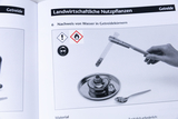 Das Bild zeigt eine Anleitung zur Durchführung eines Experiments zum Nachweis von Wasser in Getreidekörnern. Es sind Sicherheitszeichen abgebildet, und es wird ein Reagenzglas in einer Hand gehalten, während sich ein Heizgerät im Hintergrund befindet.