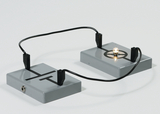 Das Bild zeigt zwei graue Kästen, die durch Kabel miteinander verbunden sind. In dem rechten Kasten leuchtet eine kleine Glühbirne, was auf einen einfachen elektrischen Schaltkreis hinweist, der für Experimente im Bildungsbereich verwendet werden kann.