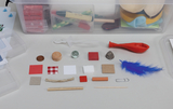 Das Bild zeigt eine Auswahl an verschiedenen Experimentiermaterialien, die für den Einsatz im Bildungsbereich vorgesehen sind. Dazu gehören unter anderem unterschiedlich farbige und strukturierte Materialien wie Holz, Stoff, Kunststoff und Metalle, angeordnet auf einem neutralen Hintergrund.